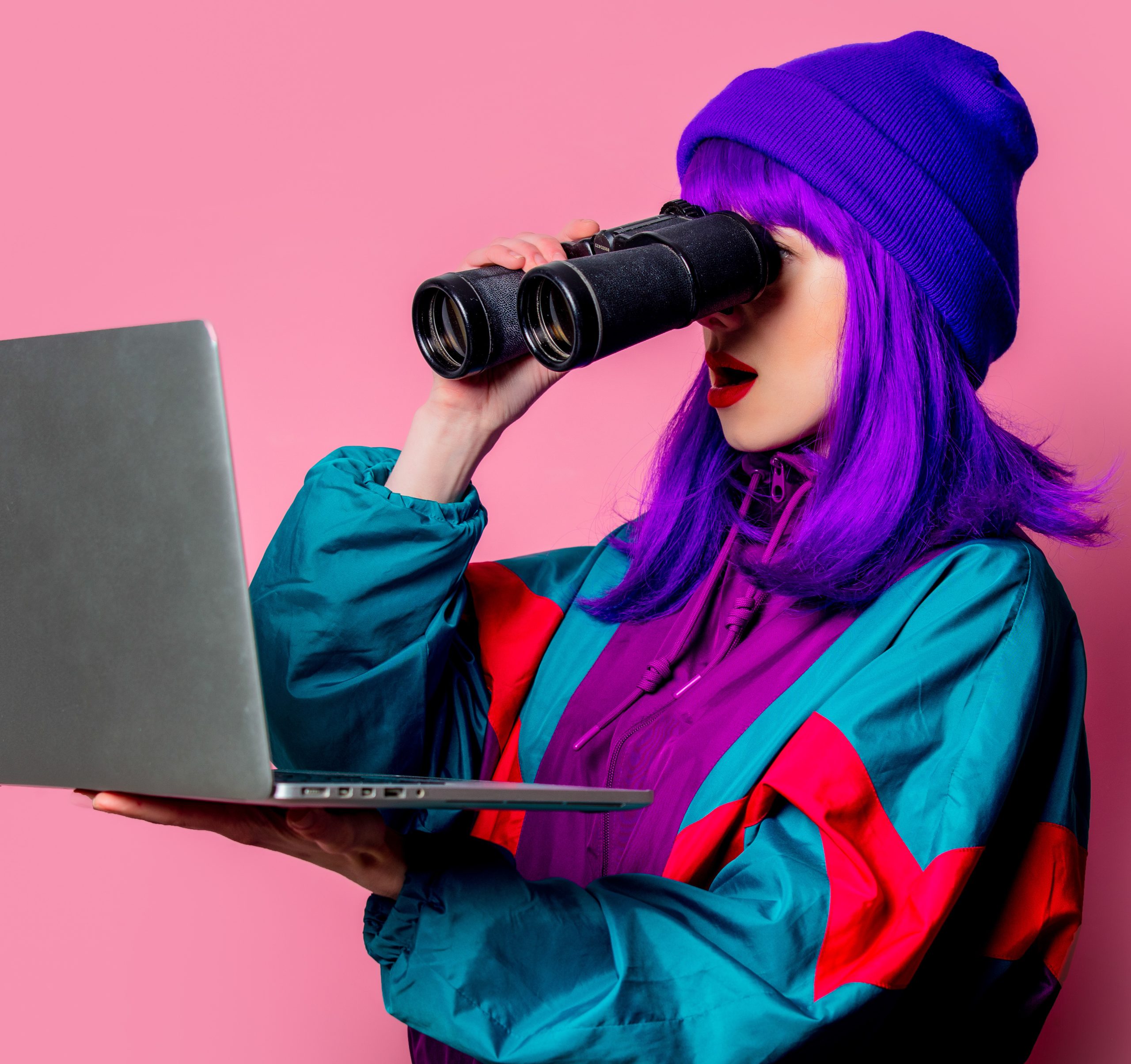 Stylish girl in 80s tracksuit watching in to laptop with binoculars on pink background looking closely at meta ads